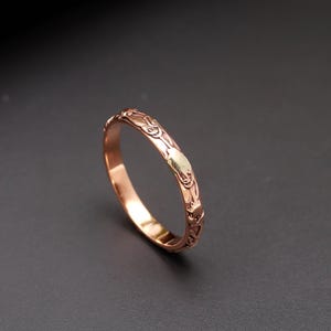 May include: A rose gold band ring featuring a continuous embossed floral pattern around the exterior surface. The metal has a polished finish that reflects light, highlighting the intricate raised details of the botanical design along the band.