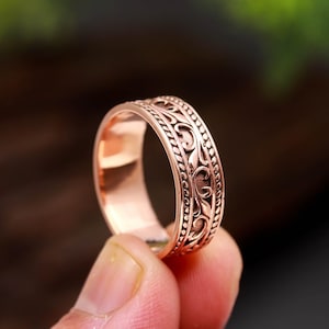 Può includere: Un anello in metallo color rame caratterizzato da una fascia centrale con un motivo a volute in rilievo, bordato su entrambi i lati da una sottile trama a perline in rilievo. L'interno della fascia è liscio e lucido con finitura metallica.