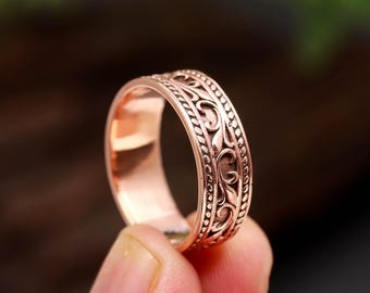 Può includere: Un anello in metallo color rame caratterizzato da una fascia centrale con un motivo a volute in rilievo, bordato su entrambi i lati da una sottile trama a perline in rilievo. L'interno della fascia è liscio e lucido con finitura metallica.