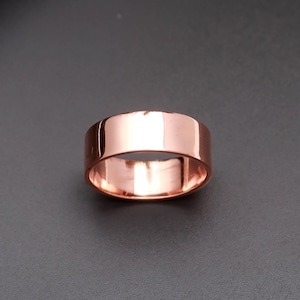 May include: A polished rose gold band ring with a smooth, flat surface and a reflective finish. The ring features a simple, wide design with a consistent width throughout the entire circumference, showcasing a warm, metallic pinkish-copper hue.
