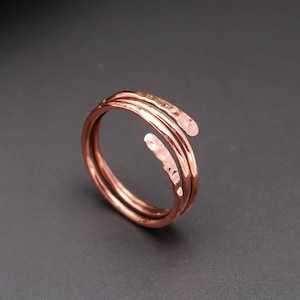 Pode incluir: Um anel de metal na cor cobre com um design de bypass e duas extremidades sobrepostas. A banda apresenta um acabamento martelado e texturizado em toda a sua superfície, criando pequenas reentrâncias ao longo da estrutura do fio.