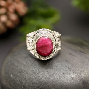 May include: A silver ring featuring a large, oval-shaped, deep pink gemstone set in the center. The metal band is decorated with intricate beaded textures and spiral patterns surrounding the stone, creating a detailed and ornate appearance.