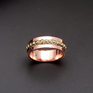Peut inclure: Une bague bicolore composée d'un large anneau poli de couleur cuivre avec une incrustation centrale texturée de couleur dorée. La bande centrale présente une surface surélevée et bosselée contrastant avec les bords lisses et plats.
