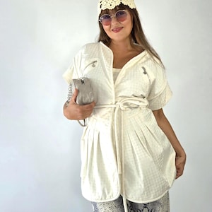 May include: A cream-colored short-sleeved jacket with a subtle grid pattern, featuring a tie-waist, two zippered chest pockets, and a V-neckline. The jacket is paired with a cream crochet beanie, round tinted sunglasses, and a small metallic silver handbag.