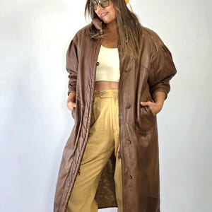 May include: A long brown leather coat with a front button closure and side pockets, worn over a cream-coloured crop top and tan trousers. A yellow beanie and oversized tinted glasses complete the ensemble, creating a layered look.