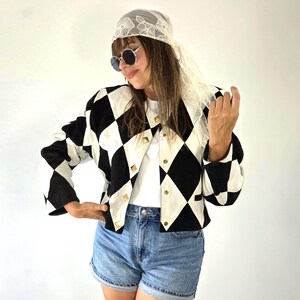 May include: A black and white diamond-patterned jacket featuring gold-toned pyramid studs along the front closure. The jacket is worn over a plain white t-shirt and blue denim shorts, paired with round sunglasses and a white floral lace headscarf.