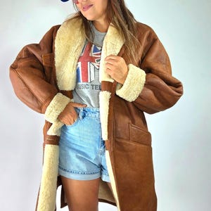 Könnte beinhalten: Ein brauner Ledermantel mit cremefarbenem Lammfellfutter, ein graues T-Shirt mit Union-Jack-Grafik und dem Text MUSIC TELEVISION, hellblaue Jeansshorts, eine blaue Baseballkappe mit weißem Logo und kniehohe, hellbraune Stiefel.