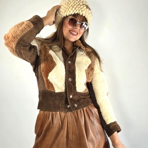 Può includere: Una giacca in camoscio marrone con inserti in pelliccia sintetica color crema, bottoni a pressione argentati e colletto. La giacca è abbinata a una gonna in pelle marrone, un berretto in maglia testurizzata color crema e occhiali da sole tondi con lenti sfumate.