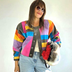 May include: A multi-colored suede jacket featuring a geometric patchwork pattern in shades of pink, yellow, orange, purple, and blue, with a dark gray front placket, round buttons, and a white shirt underneath, paired with light blue denim jeans.