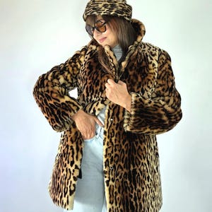 May include: A leopard print faux fur coat with a matching beanie hat, worn over a light gray turtleneck sweater and light blue denim jeans. The coat features a high collar and a button-front closure, showcasing a classic brown and black animal pattern.