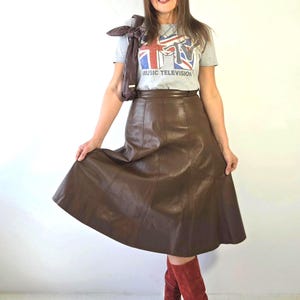Puede incluir: Una camiseta gris de manga corta con un gráfico de la bandera Union Jack y el texto MTV MUSIC TELEVISION, combinada con una falda midi de cuero marrón oscuro con costuras verticales y un bolso de hombro de cuero marrón oscuro a juego.