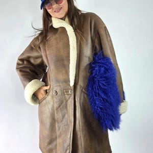 Puede incluir: Un abrigo de cuero marrón con forro de borreguillo color crema, que presenta un gran acento de piel sintética azul vibrante en el lado izquierdo, una gorra de béisbol azul con letras LA blancas y gafas de sol tintadas.
