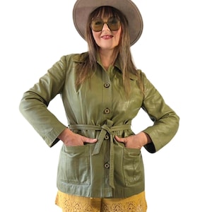 Pode incluir: Uma jaqueta de couro verde-oliva com gola, fecho frontal de botões, mangas compridas e um cinto de amarrar a condizer. A jaqueta inclui dois bolsos frontais grandes e é combinada com calções amarelos com um detalhe de bainha em renda.