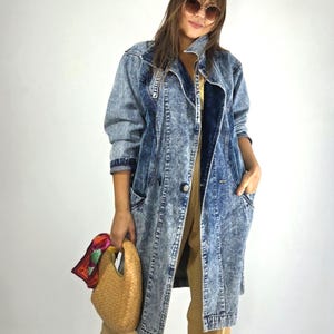 Peut inclure: Un long manteau en jean bleu délavé à l'acide avec un col cranté, une fermeture à un bouton sur le devant et deux poches latérales. Le manteau est porté sur un pantalon beige et accompagné d'un sac à main tressé beige avec un foulard coloré.