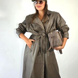 May include: An olive green baseball cap with a white NY logo, round sunglasses with thin frames, and a long, taupe-coloured leather trench coat featuring a wide collar, long sleeves, and a matching tie belt at the waist, paired with a taupe leather handbag.