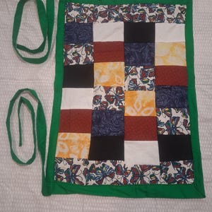 May include: A rectangular fabric apron featuring a patchwork design of various squares in solid black, white, red, and yellow, alongside patterned squares with butterfly motifs. The apron is bordered by a solid green trim and includes two long green ties.