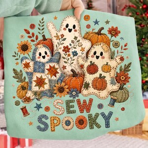 Op de afbeelding: Een lichtgroen sweatshirt met een afbeelding van drie lachende spoken omringd door pompoenen, zonnebloemen en naaiknoppen. De tekst SEW SPOOKY is gedrukt in een stiksel-stijl lettertype. Het ontwerp bevat quiltpatronen en garenklosjes.