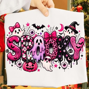 以下が含まれることがあります： 大きなスタイリッシュな文字で「SPOOKY」と書かれたカラフルなグラフィックがプリントされた白いTシャツ。ピンクと黒のゴースト、カボチャ、コウモリ、クモの巣、星、目玉、リボンが描かれた、遊び心のあるハロウィーンをテーマにしたデザインです。