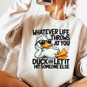 Puede incluir: Una sudadera de cuello redondo color crema con el gráfico de un pato blanco con gafas de sol negras, apoyando la cabeza en su ala. El texto dice: WHATEVER LIFE THROWS AT YOU DUCK AND LET IT HIT SOMEONE ELSE en letras negras negritas.