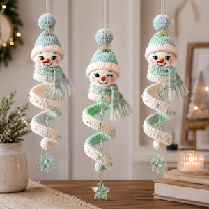 May include: Three hanging crochet snowman ornaments featuring mint green and white yarn, each wearing a matching beanie with a pom-pom and a scarf. The bodies are spiraled, accented with small green beads, and finished with a hanging crystal star.