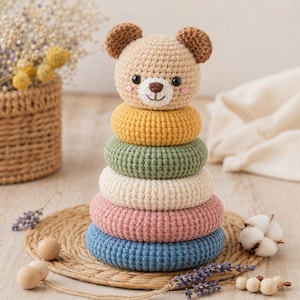 May include: A crochet stacking toy featuring a beige bear head topper with brown ears and black eyes, sitting atop five stacked rings in yellow, sage green, cream, dusty pink, and blue. The toy is constructed from yarn with a visible stitch texture.