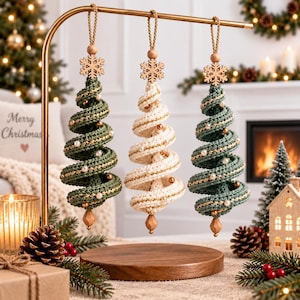May include: Three spiral Christmas tree ornaments hanging from a gold metal stand. Two ornaments are dark green and one is cream, all featuring crocheted textures, gold metallic thread accents, small white pearls, and a wooden snowflake topper.
