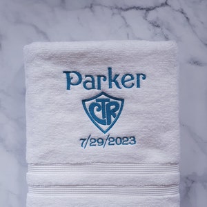 May include: White embroidered towel with the name "Parker", a shield logo, and the date 7/29/2023 in teal. The towel has a textured surface and horizontal stripes near the bottom.