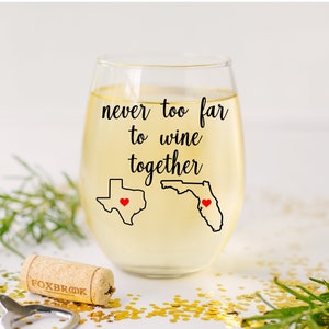 May include: A stemless wine glass filled with white wine, featuring the text "never too far to wine together." The glass has black outlines of the states of Texas and Florida, each with a red heart. A cork and bottle opener are in the foreground.