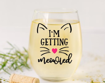 Puede incluir: Una copa de vino sin pie llena de un líquido claro, con un diseño de cara de gato negro y la frase "I'M GETTING meowied" en letras negras con un corazón rosa. Un corcho y un abridor de botellas están cerca.