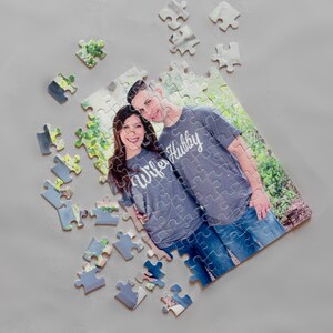 May include: A personalized jigsaw puzzle featuring a couple wearing matching gray t-shirts with the words "Wife" and "Hubby" printed on them. The puzzle pieces are scattered on a gray surface, with a photo of the couple in the center.