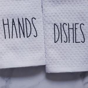 May include: Two white kitchen towels with black embroidery. The towel on the left says "HANDS" and the towel on the right says "DISHES".