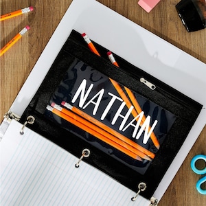 May include: Black zippered pencil pouch with a clear plastic window that says "NATHAN" in white lettering. The pouch is open and has several yellow pencils inside.