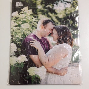 May include: A rectangular jigsaw puzzle featuring a couple embracing, set against a backdrop of white flowers and greenery. The puzzle is packaged in clear plastic. The man wears a purple shirt, and the woman wears a white dress.