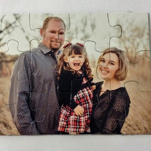 May include: A family portrait printed on a jigsaw puzzle. The image shows a man, a woman, and a young girl smiling. The girl wears a red and black plaid dress. The man wears a gray plaid shirt. The woman wears a black top.