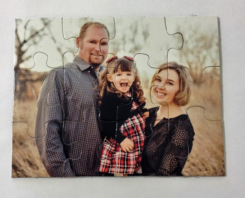 May include: A family portrait printed on a jigsaw puzzle. The image shows a man, a woman, and a young girl smiling. The girl wears a red and black plaid dress. The man wears a gray plaid shirt. The woman wears a black top.