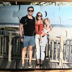 May include: A family portrait puzzle featuring a man, a woman, and a child on a beach boardwalk. The puzzle pieces are interlocked, displaying a sunny day scene with the ocean in the background. The family is wearing sunglasses.