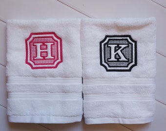 May include: Two white towels with embroidered letters. The towel on the left has a pink "H" in a black frame. The towel on the right has a black "K" in a white frame.