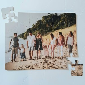 May include: A custom photo puzzle featuring a family walking on a beach. The puzzle is mostly assembled, with a few pieces separate. The image shows a group of people, including children, holding hands and walking along the shoreline. The puzzle is on a white surface.