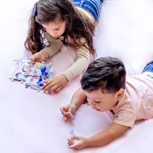 May include: Two children are lying on a pink surface, assembling a jigsaw puzzle. The puzzle pieces feature a colorful image. One child is holding puzzle pieces, while the other is reaching for a piece. The children are wearing casual clothing.