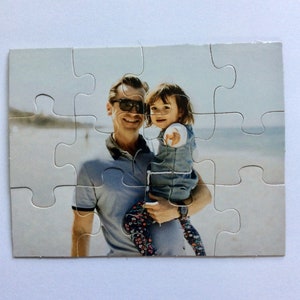 May include: A completed jigsaw puzzle featuring a man holding a young child on a beach. The man wears sunglasses and a gray polo shirt. The child points with one hand and wears a denim vest and patterned pants. The background shows a blurred beach scene.