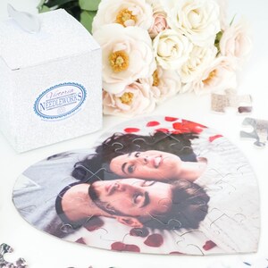 May include: A heart-shaped jigsaw puzzle featuring a couple's photo, surrounded by red rose petals. A white glitter gift box with the text "Victoria Needleworks" and a bouquet of white and pink roses are also present.