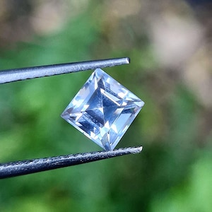Puede incluir: Una piedra preciosa transparente e incolora de corte princesa sostenida por unas pinzas de metal. La piedra de forma cuadrada presenta múltiples facetas triangulares que reflejan la luz, creando un aspecto cristalino y claro.