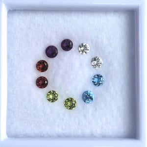 May include: A collection of nine round, faceted gemstones arranged in a circular pattern inside a white display box. The stones feature a spectrum of colors including purple, white, blue, light green, and deep red, each showing distinct light reflection.