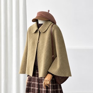 May include: A tan textured cape jacket with a rounded collar and front buttons, worn over a dark brown turtleneck and a brown plaid pleated skirt. A brown beret and a brown shoulder bag with a thin strap complete the ensemble.
