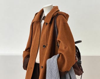May include: A hooded, rust-coloured coat with dark buttons, worn over a white jumper and brown trousers. A grey ribbed scarf is draped over the arm, and a brown backpack with dark straps is positioned on the back of the coat.