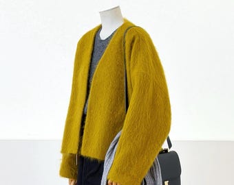May include: A fuzzy mustard yellow open-front cardigan worn over a grey sweater and black skirt. A grey ribbed scarf hangs over the shoulder, and a black shoulder bag with a gold-tone metal H-shaped clasp is carried on the side.