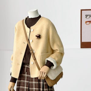 May include: A cream-colored, long-sleeved, button-down jacket with a round neckline, worn over a dark brown sweater and a brown plaid pleated skirt. A tan crossbody bag with a white faux-fur flap is worn over the shoulder.