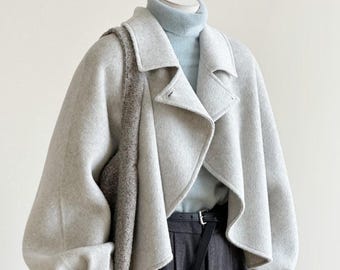 May include: A light grey cropped wool-blend jacket with wide sleeves and a notched collar, worn over a light blue turtleneck sweater. The outfit includes dark charcoal grey pleated trousers with a black belt and a large, textured brown shoulder bag.