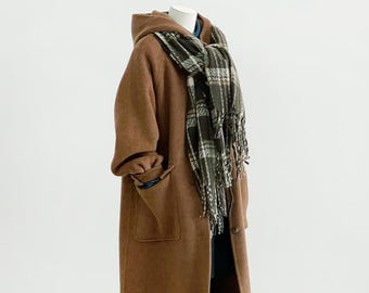 May include: A long, brown, hooded coat with large patch pockets, worn with a dark green and white plaid scarf featuring fringe edges. The coat is displayed on a mannequin, paired with black leggings and brown leather buckled shoes.