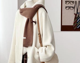 May include: A cream-coloured hooded coat with long sleeves and two front pockets, worn with a brown knit scarf featuring a small white bow detail. A brown crossbody bag with a textured cream flap and a brown plaid skirt are also visible.